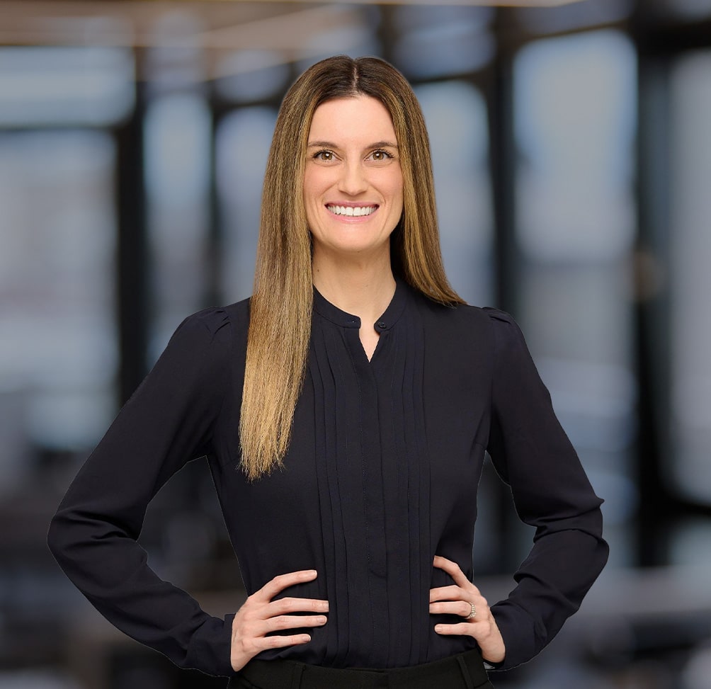 Professional woman with long straight hair smiling, standing with hands on hips in a dark blouse, blurred office background