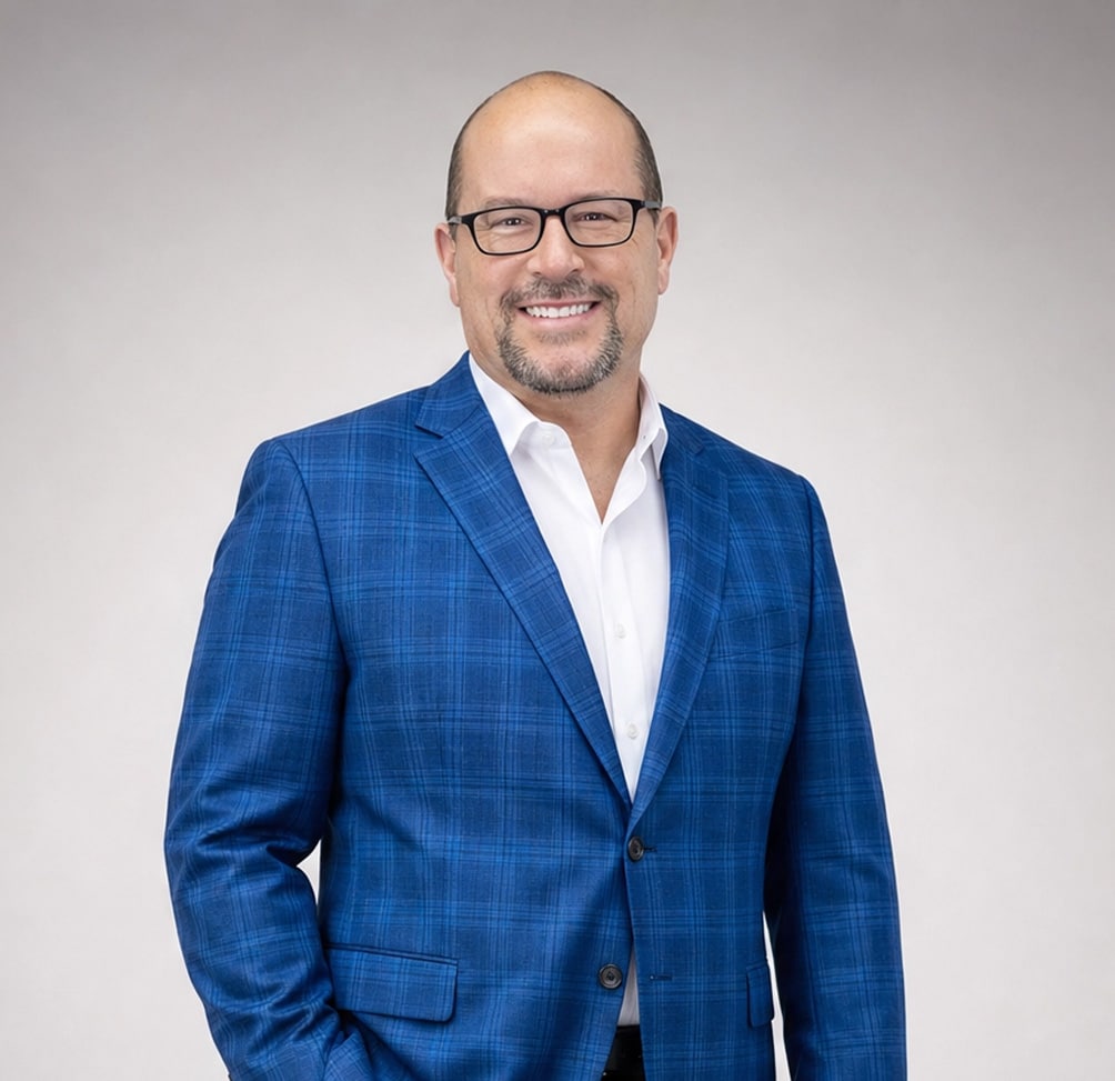 Professional headshot of a smiling man wearing a blue plaid blazer and white shirt against a light gray background.
