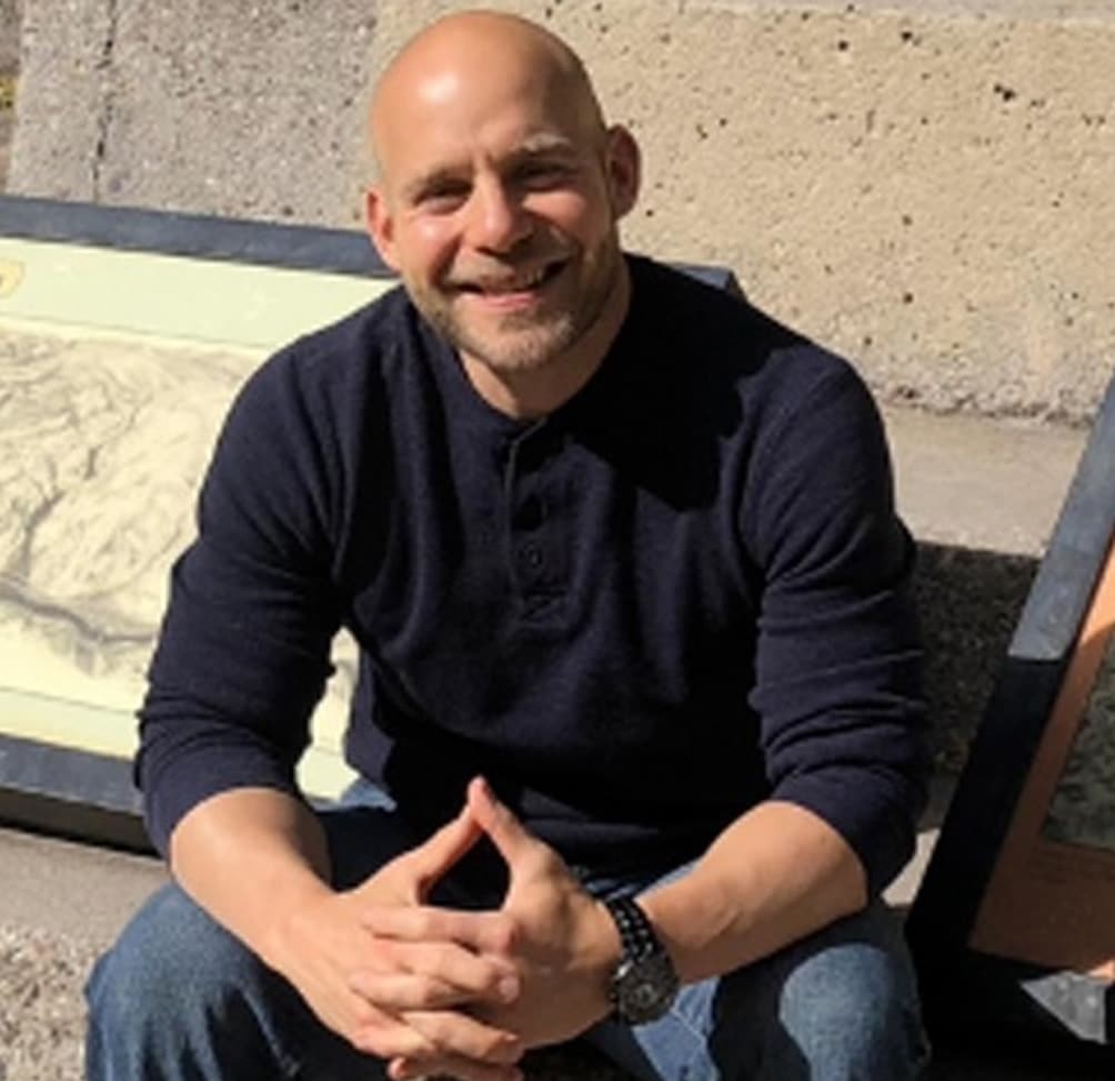 Smiling bald man in a navy sweater sits on a bench outdoors, hands clasped in his lap.
