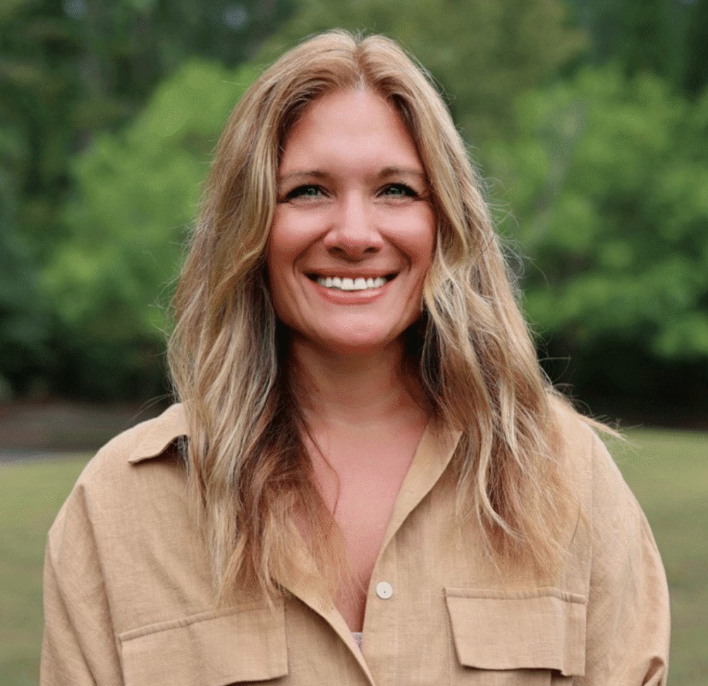 Smiling woman with long blond hair outdoors, wearing a beige button-up shirt.