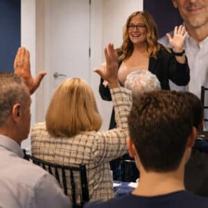 Woman presenter standing at the front, smiling, with hands raised to engage a seated audience in a meeting or workshop session.