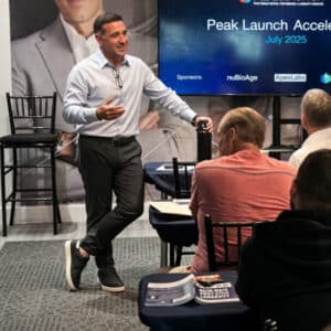 Speaker in a light blue shirt stands and gesticulates to an audience at a conference, with a slide reading 'Peak Launch Accelerate July 2025' behind him.