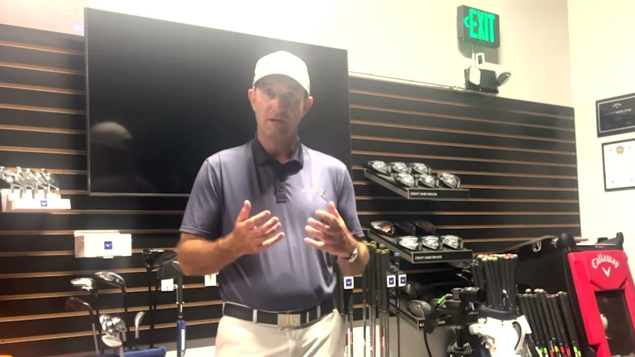 Man in a polo shirt and cap speaking to camera in a golf equipment showroom with clubs on display behind him.