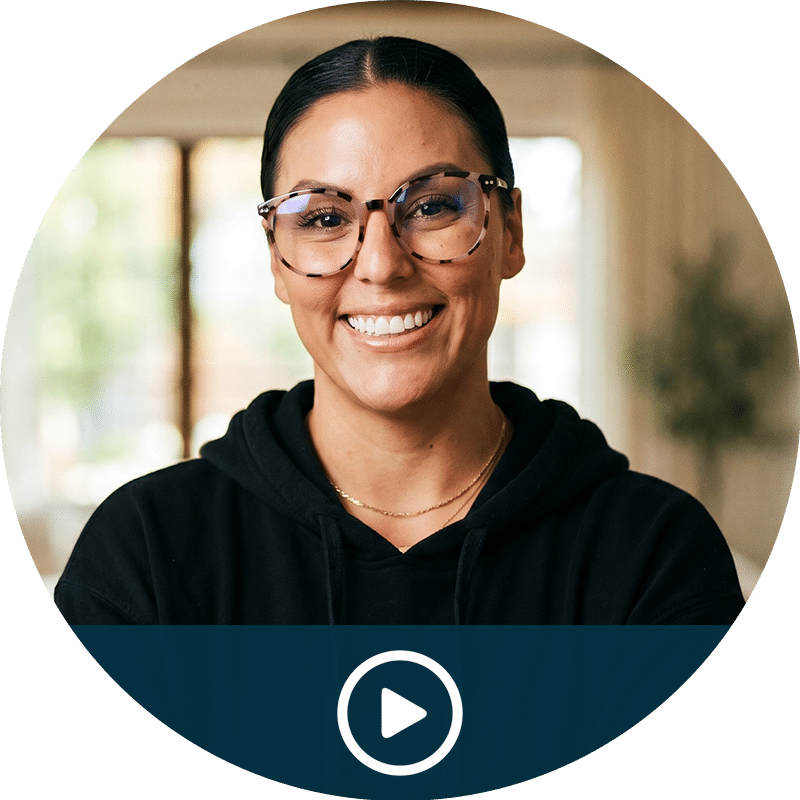 Video thumbnail: smiling woman with glasses in a black hoodie, indoors, with a play button overlay (ready to play).