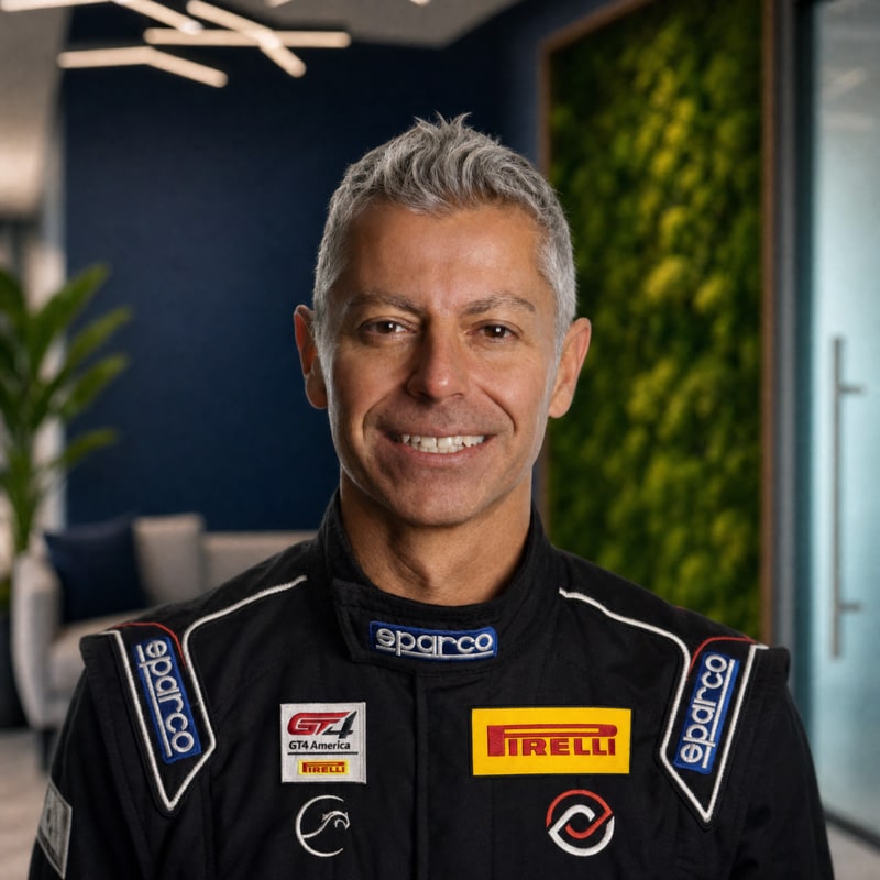 Smiling male race car driver in a black Sparco suit with Pirelli and GT4 America logos, indoors.