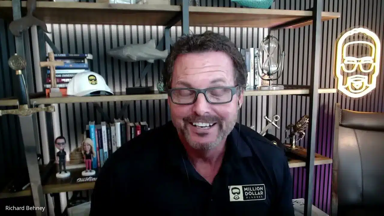 Man in a dark shirt and glasses smiling during a video call in a modern office with shelves, books, and a neon logo in the background.