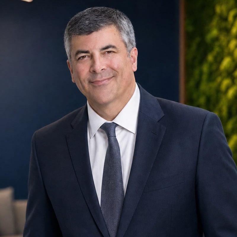 Smiling middle-aged man in a navy suit and tie, standing in a professional head-and-shoulders portrait indoors.
