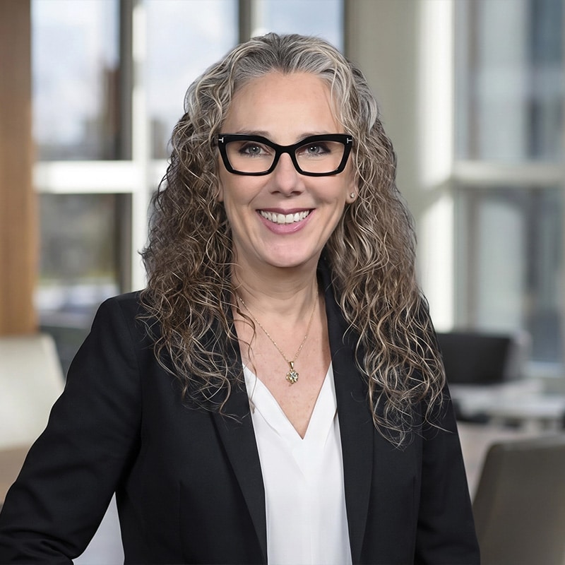 Professional woman with curly gray hair and black-framed glasses, smiling in a modern office setting