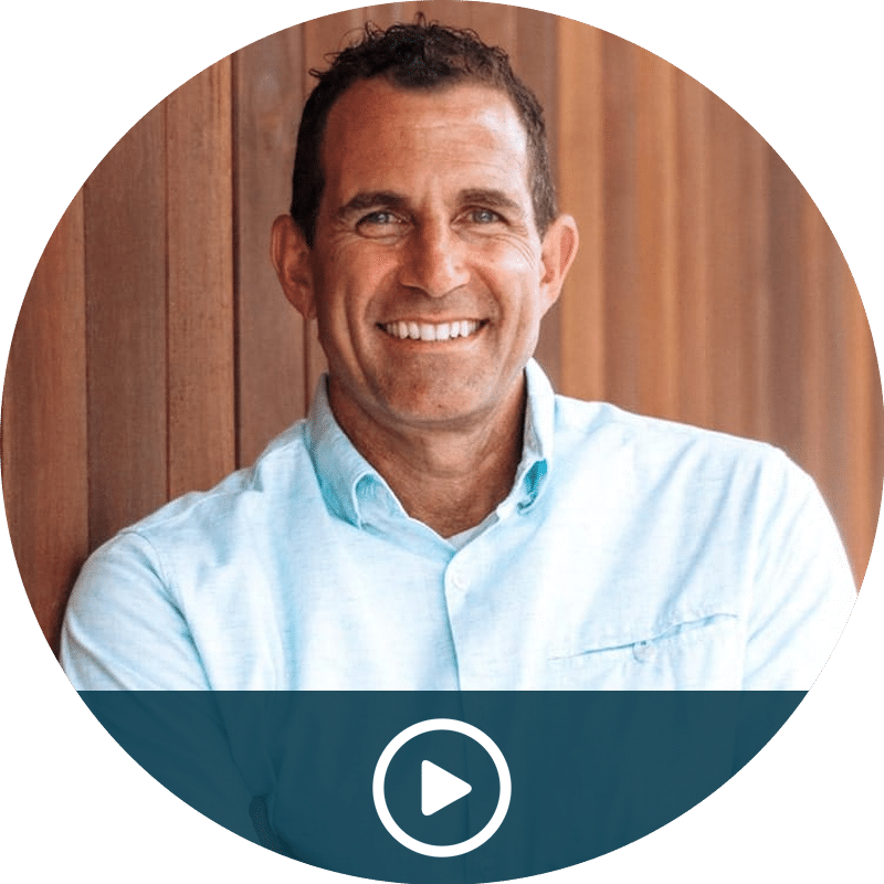 Video thumbnail: smiling man in a light blue shirt against wood panel background, with a play button overlay